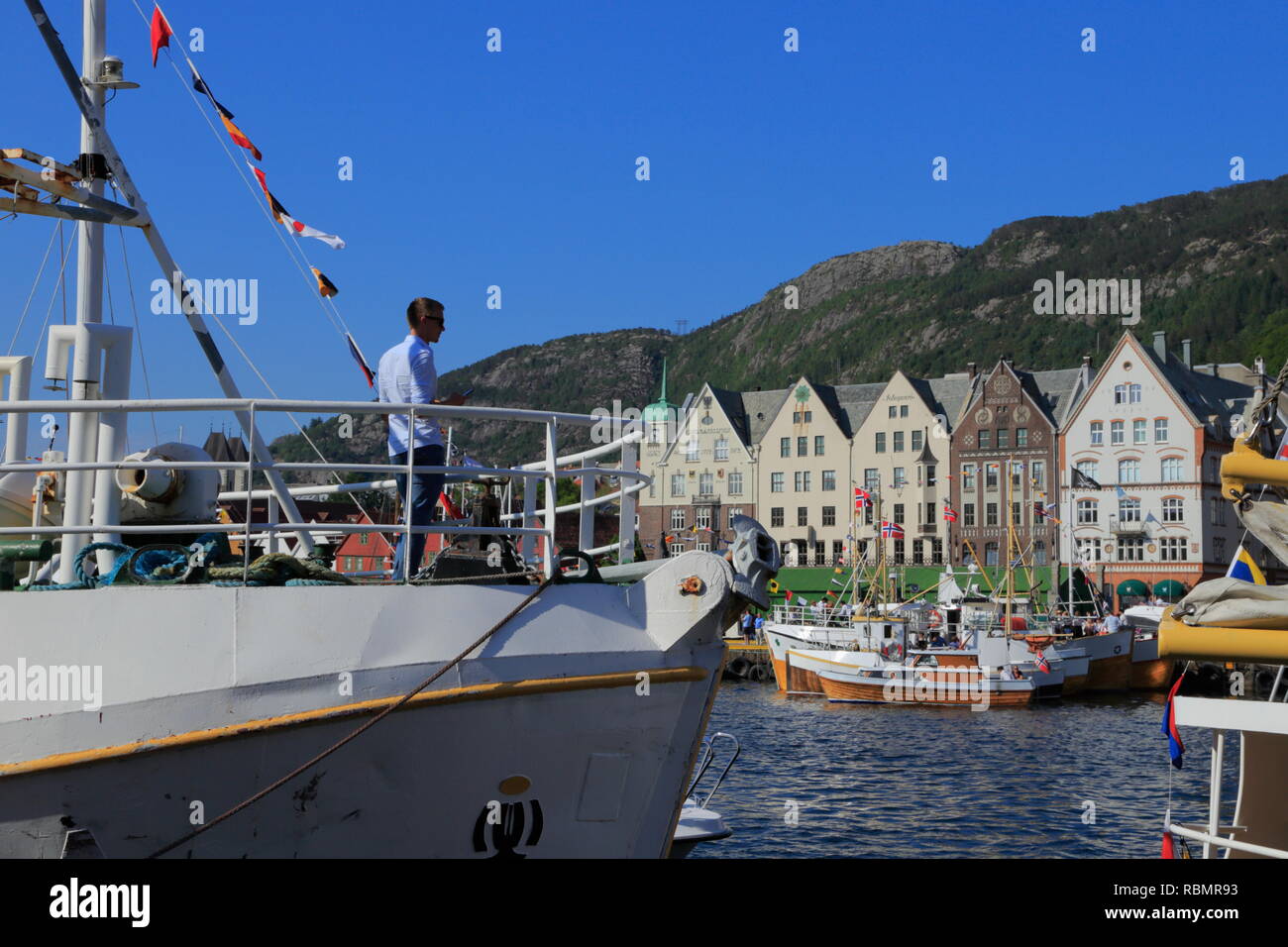 A young man stands on the deck of a boat docked in Bergen harbour, Norway, with a view of the Hanseatic buildings along Bryggen. Stock Photo