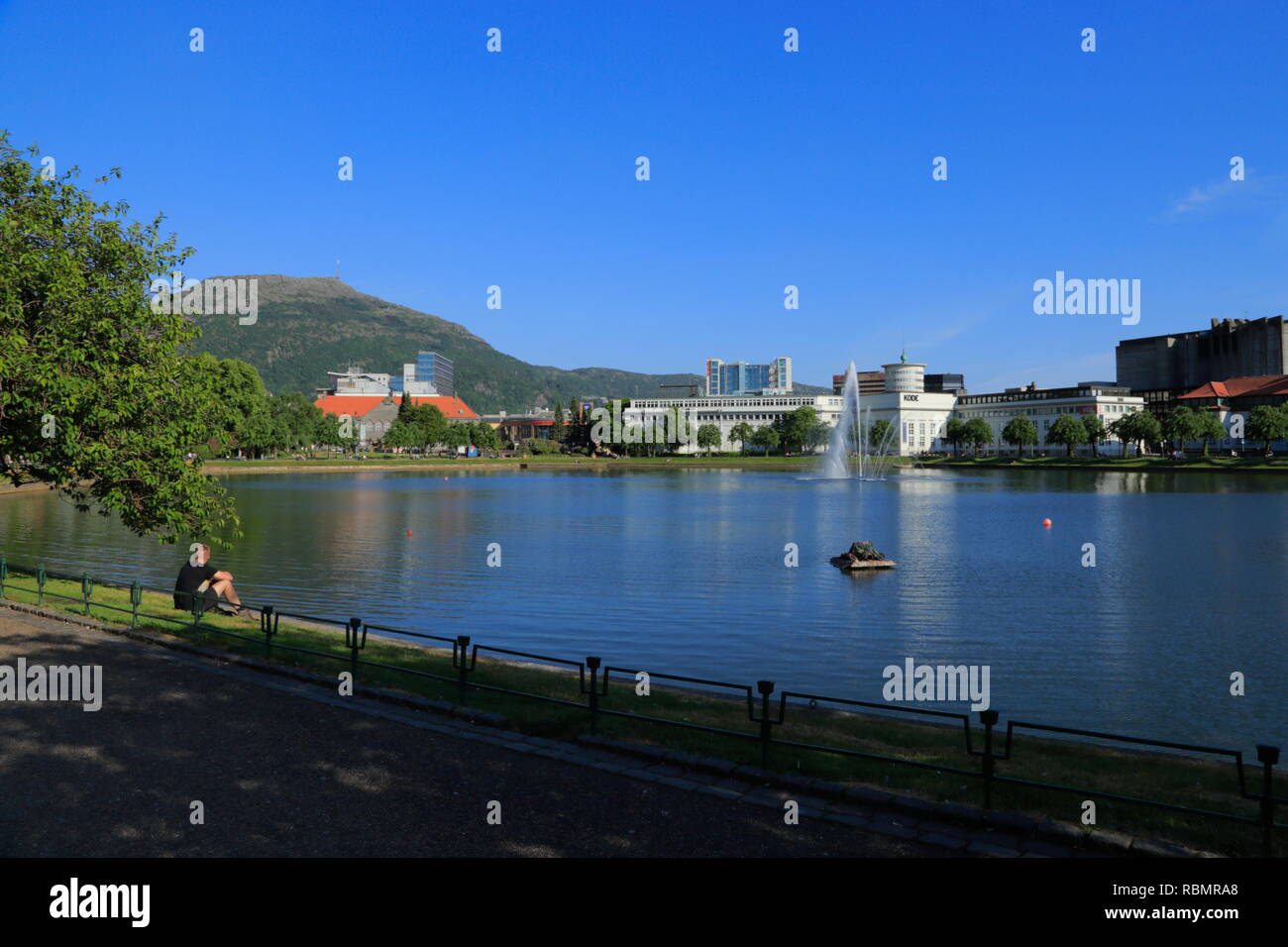 A man sits on the bank of the lake, Lille Lungegårdsvannet, in Bergen city, Norway, with a view of Mount Ulriken, KODE 4 and other buildings. Stock Photo