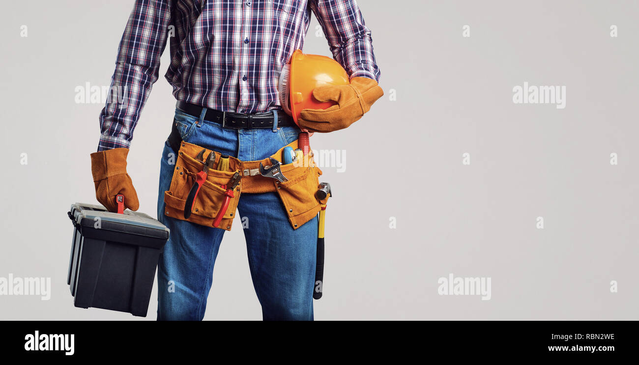 Faceless man in plaid shirt and gloves wearing utility belt and holding box of instruments and hardhat on gray backdrop Stock Photo