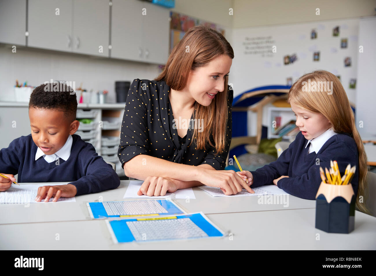 Female school teacher sitting between two primary school kids at a table in a classroom, helping a girl with her work, close up Stock Photo