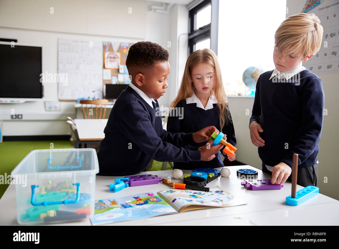 A girl and two boys standing at a table in a primary school classroom working together with toy construction blocks, close up Stock Photo