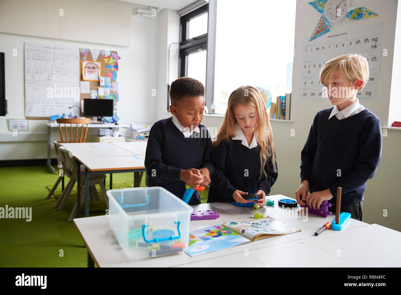 A girl and two boys standing at a table in a primary school classroom working together with toy construction blocks Stock Photo