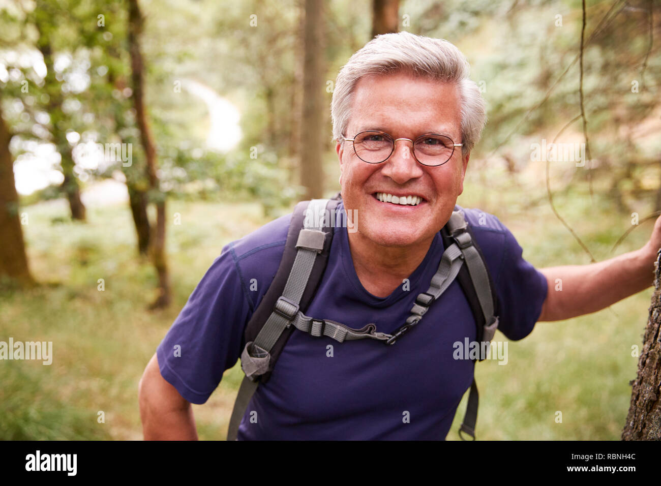 Middle aged Caucasian man taking a break leaning on a tree during a hike in a forest, smiling to camera, close up Stock Photo