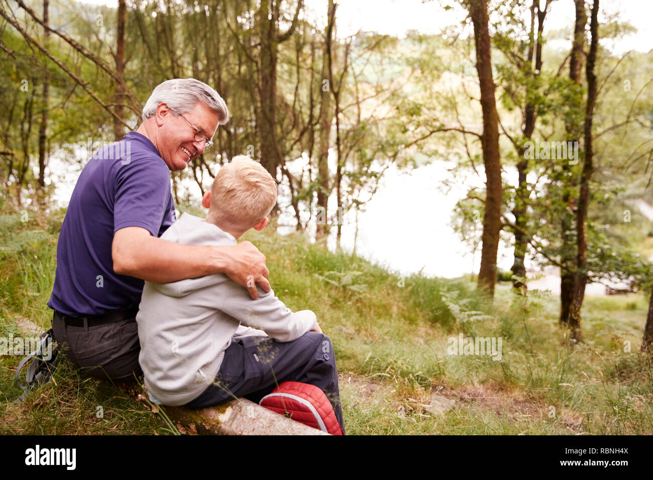 Grandfather and grandson on a hike sitting on a fallen tree in a forest, looking at each other, back view Stock Photo