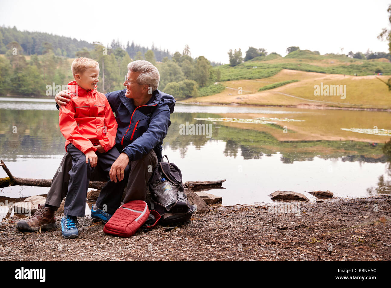 Grandson sitting on his grandfather's knee at the shore of a lake smiling, Lake District, UK Stock Photo