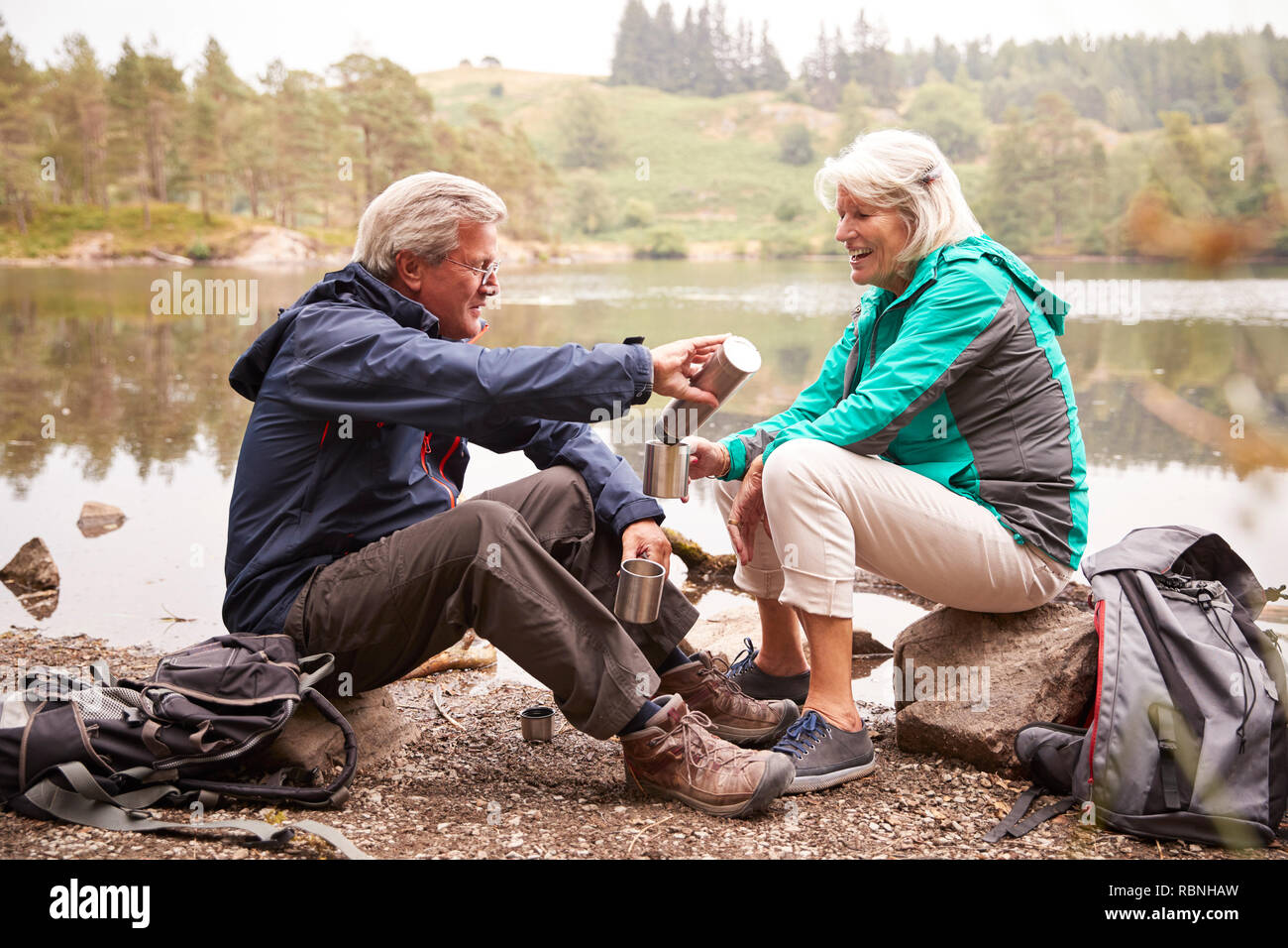 Senior couple sitting by a lake pouring coffee from a flask during a camping holiday, Lake District, UK Stock Photo
