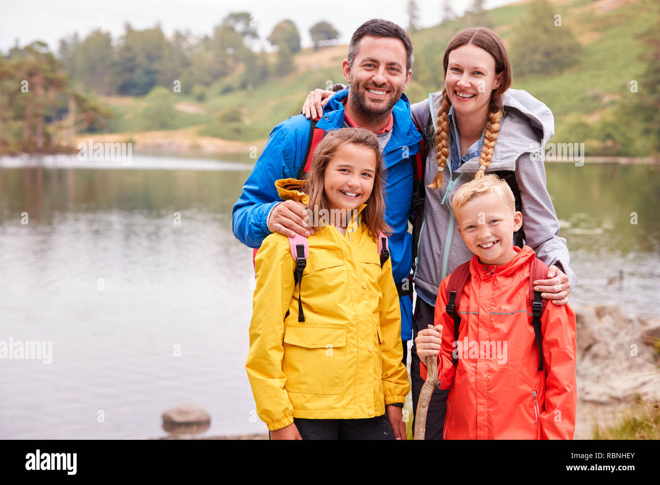 Young family standing on the shore of a lake in the countryside looking to camera smiling, Lake District, UK Stock Photo