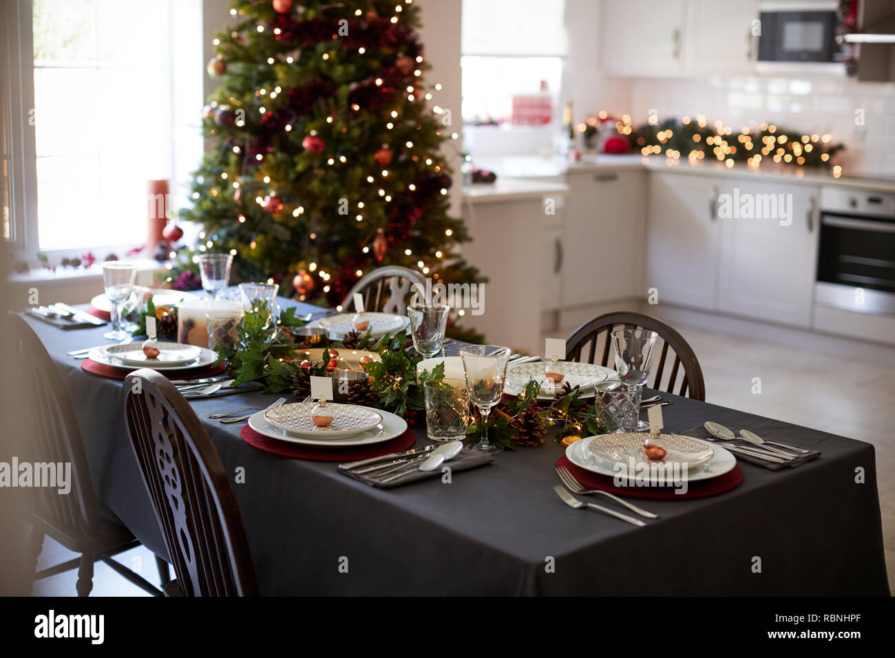 Christmas table setting with bauble name card holders arranged on plates and green and red decorations with Christmas tree and kitchen in the background Stock Photo