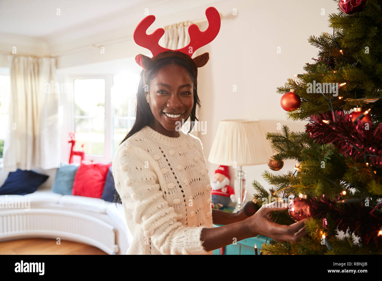 Portrait Of Woman Wearing Antlers Hanging Decorations On Christmas Tree At Home Stock Photo