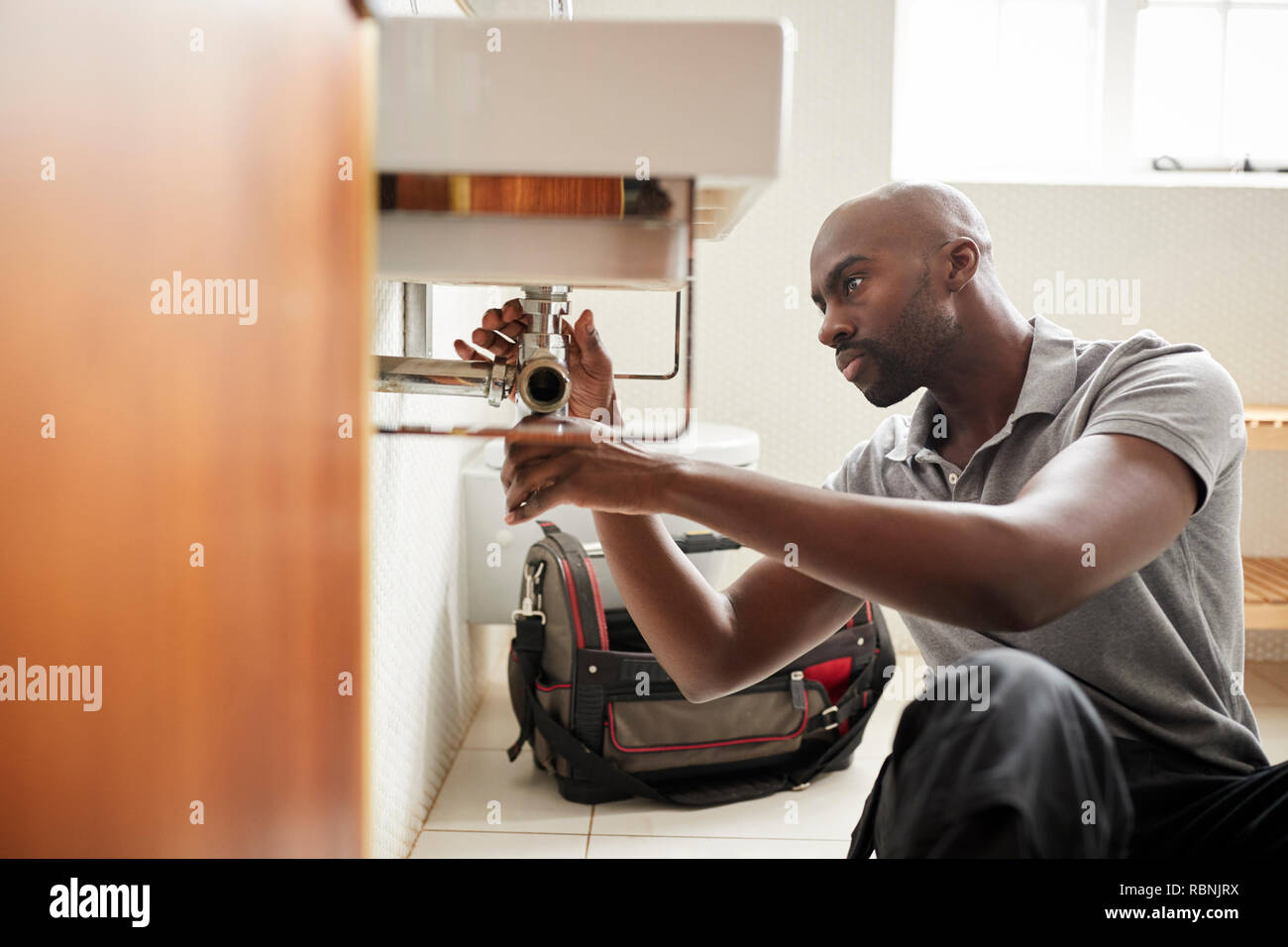 Young black male plumber sitting on the floor fixing a bathroom sink, seen from doorway Stock Photo