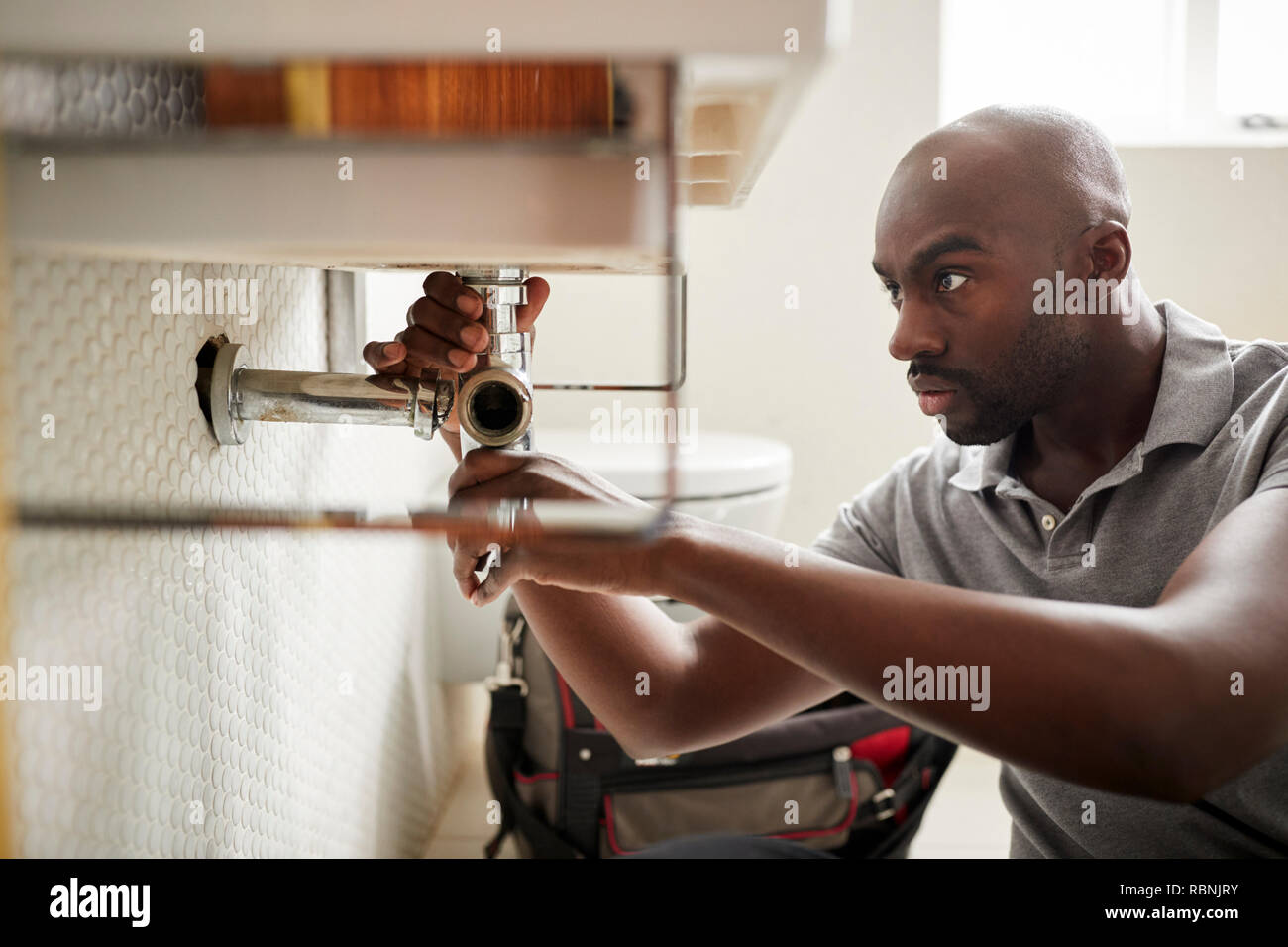 Young black male plumber sitting on the floor fixing a bathroom sink, close up Stock Photo