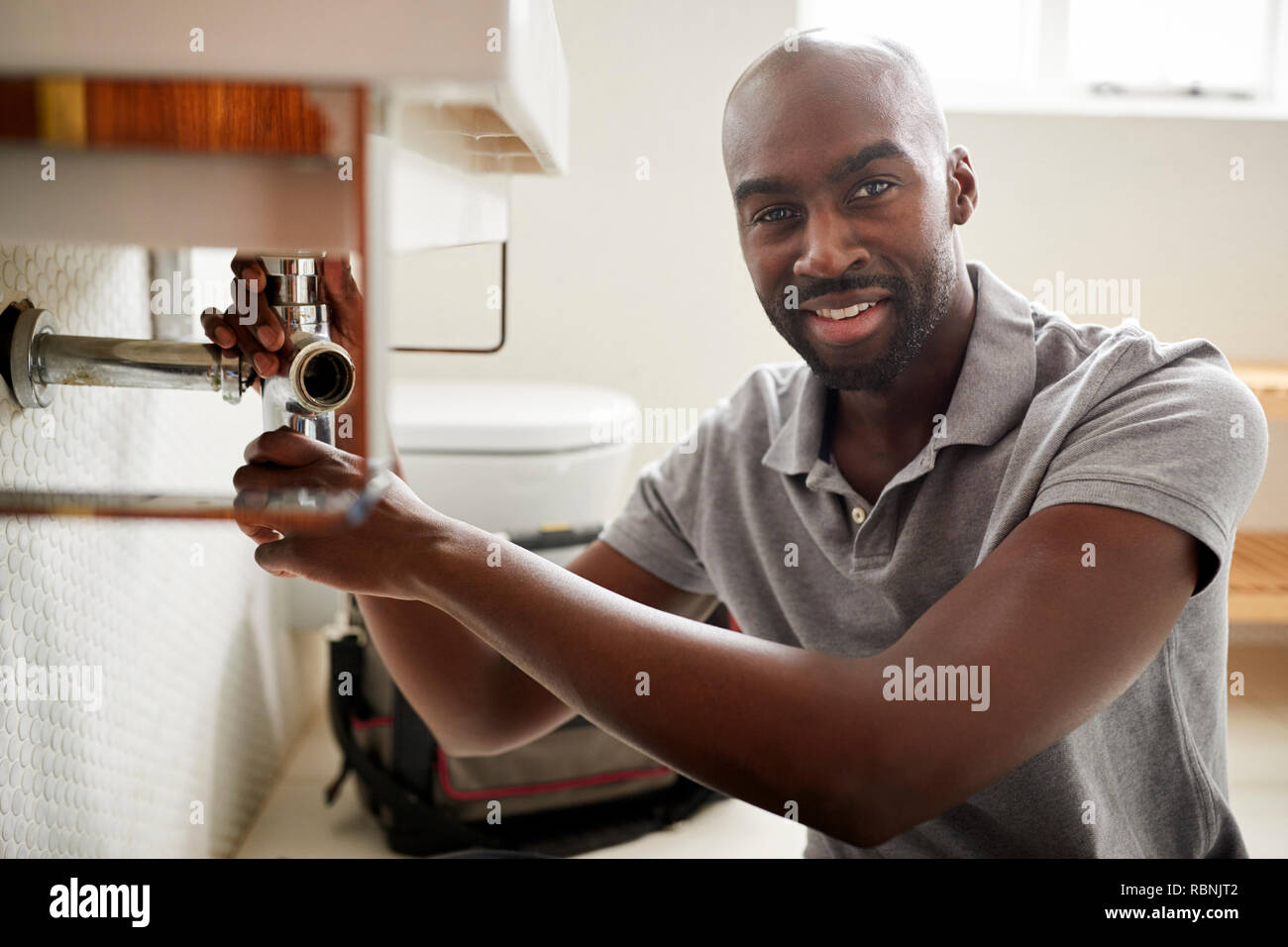 Young black male plumber sitting on the floor fixing a bathroom sink, looking to camera, close up Stock Photo