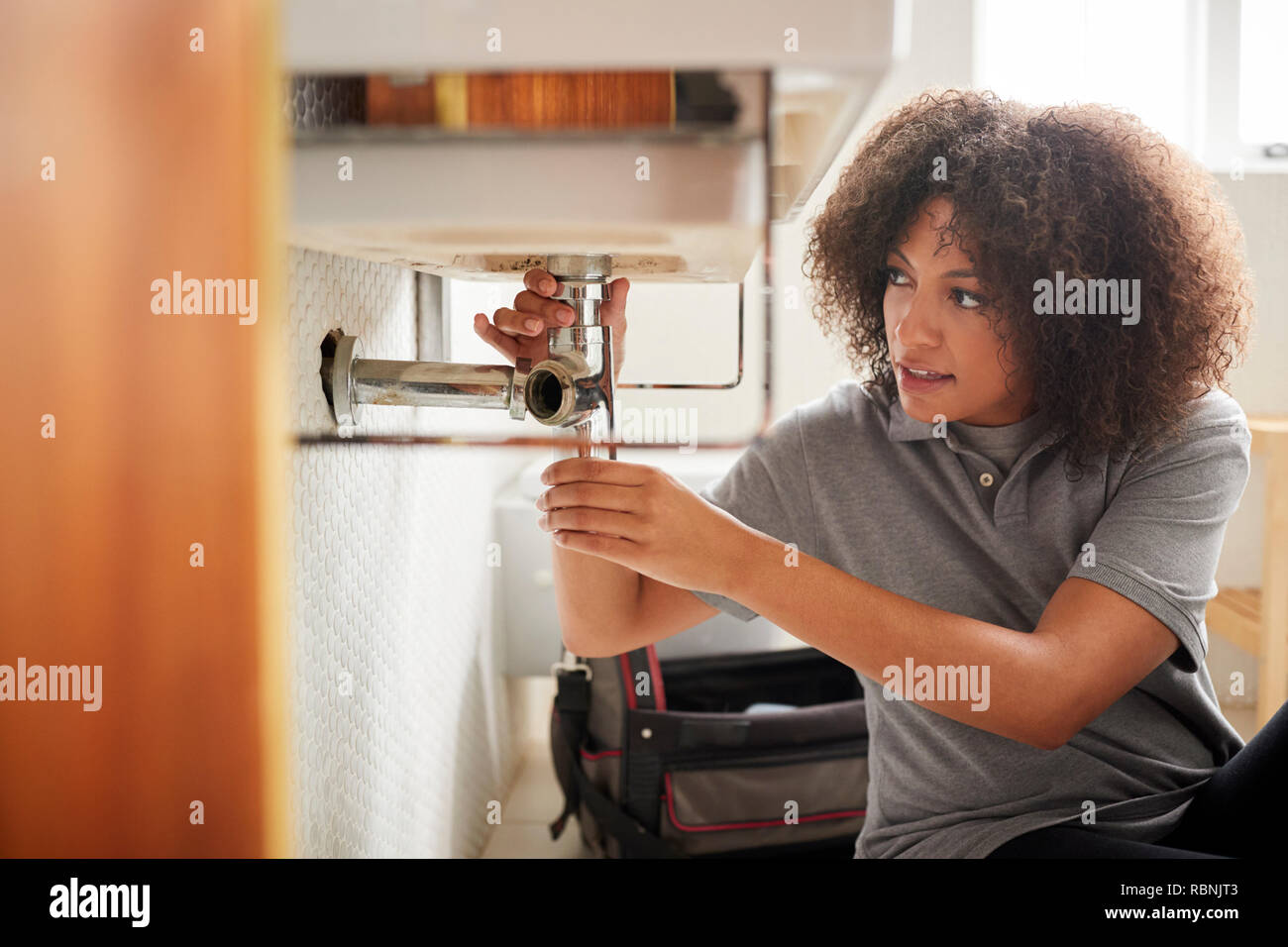 Young black female plumber sitting on the floor fixing a bathroom sink, seen from doorway Stock Photo