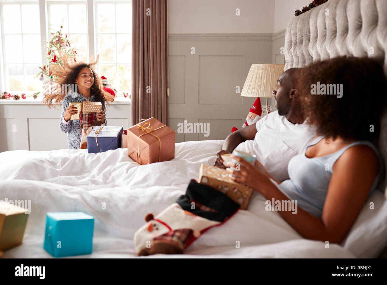 Young girl in her parents’ bedroom carrying presents on Christmas morning, parents sitting up in bed, side view Stock Photo
