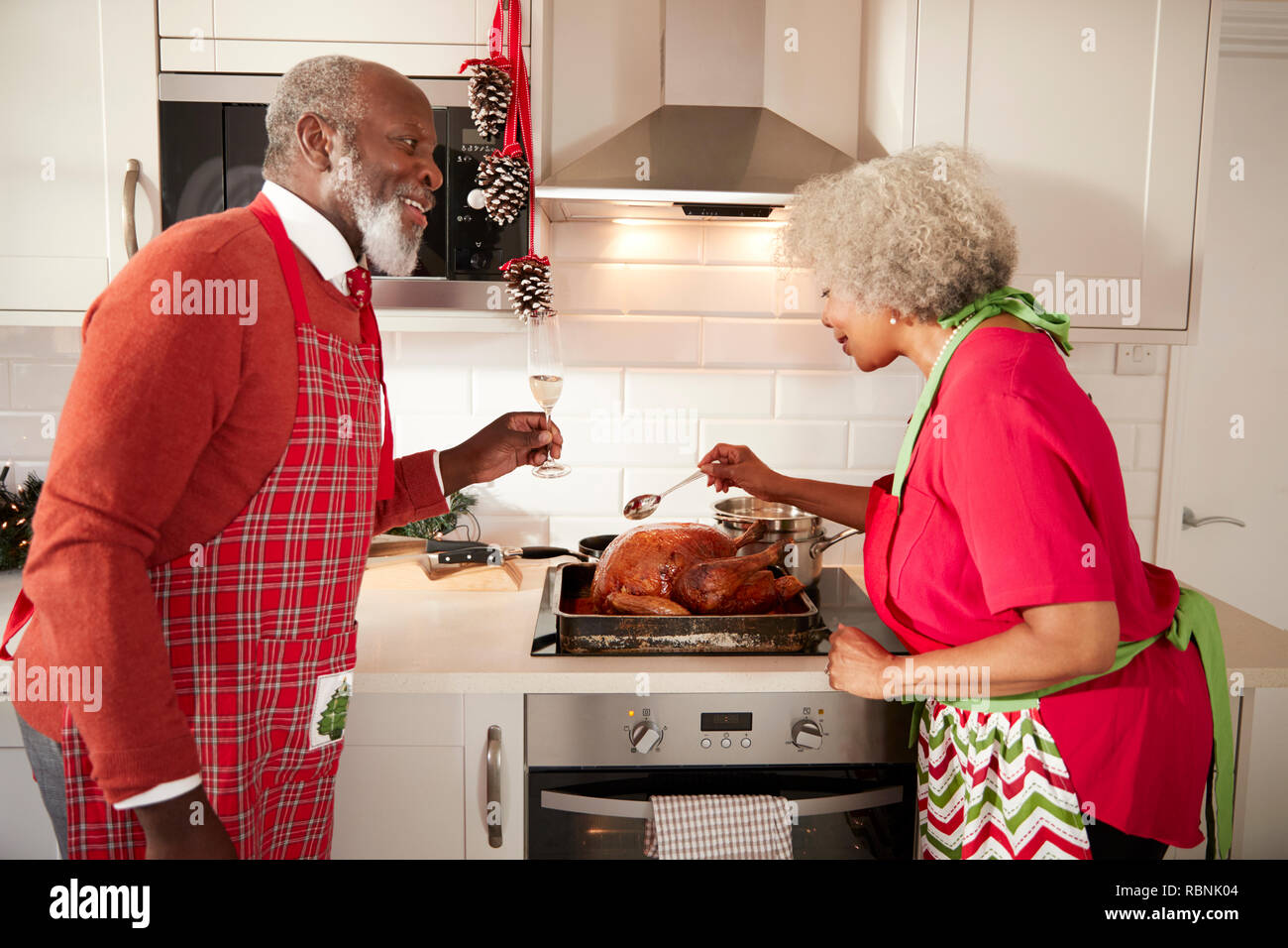 Mature black couple preparing Christmas dinner in their kitchen, man raising a glass as his wife bastes the roast turkey Stock Photo