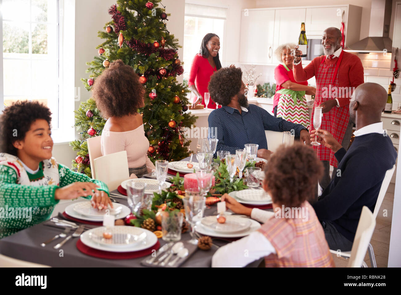 Mixed race, multi generation family gathering in dining room to sit down for Christmas dinner, grandfather holding champagne bottle Stock Photo