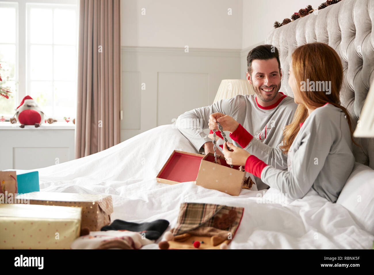 Woman Opening Gift Of Necklace In Bed At Home As Couple Exchange Presents On Christmas Day Stock Photo