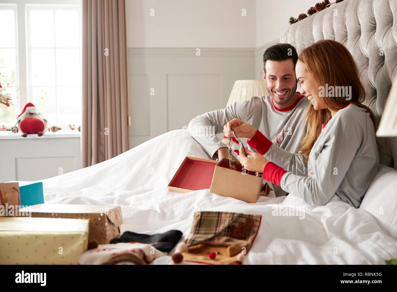 Woman Opening Gift Of Necklace In Bed At Home As Couple Exchange Presents On Christmas Day Stock Photo