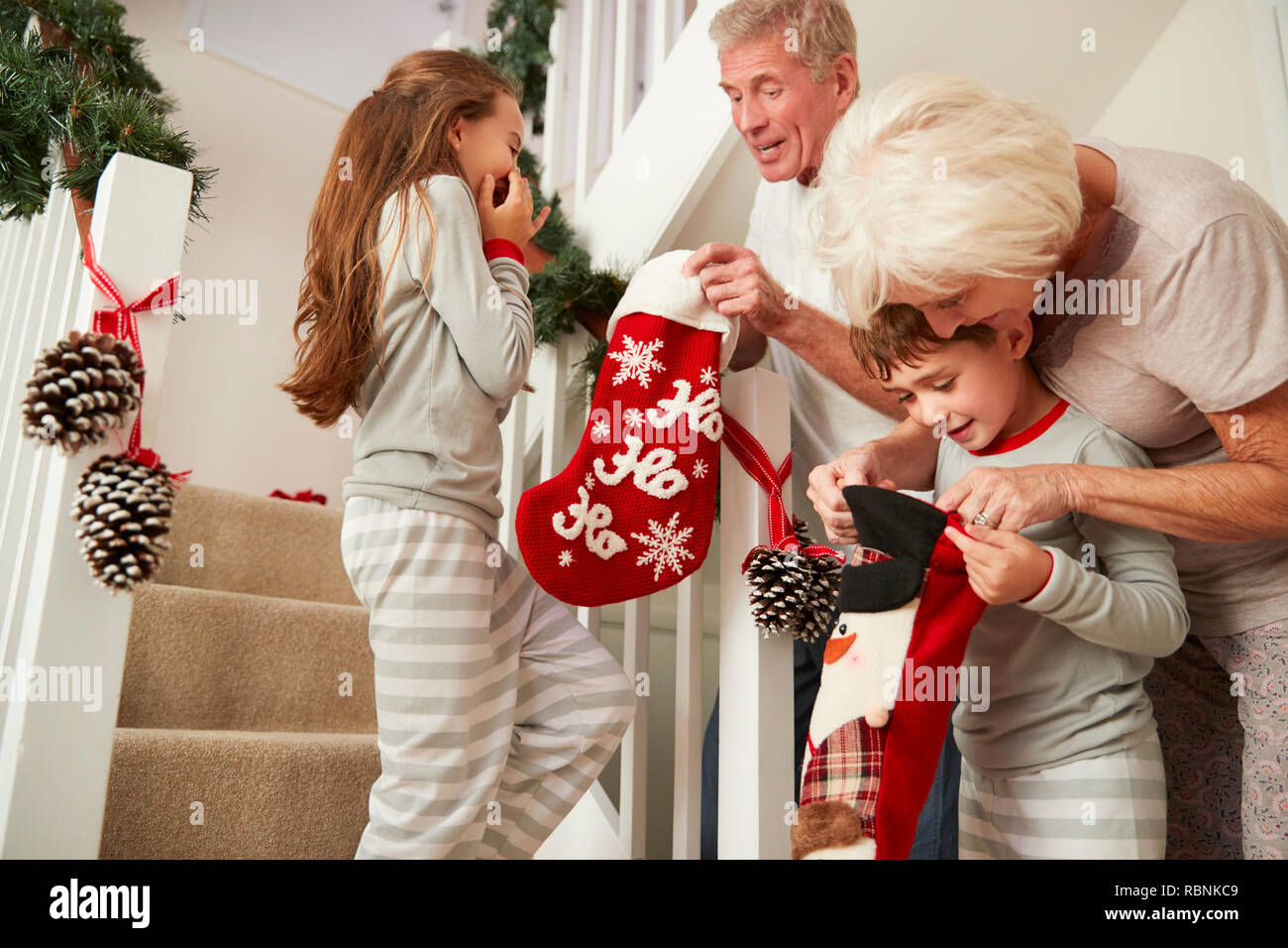 Grandparents Greeting Excited Grandchildren Wearing Pajamas Running Down Stairs Holding Stockings On Christmas Morning Stock Photo