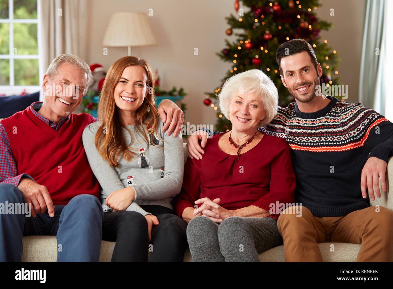 Portrait Of Senior Parents With Adult Offspring Wearing Festive Jumpers Sitting On Sofa In Lounge At Home On Christmas Day Stock Photo