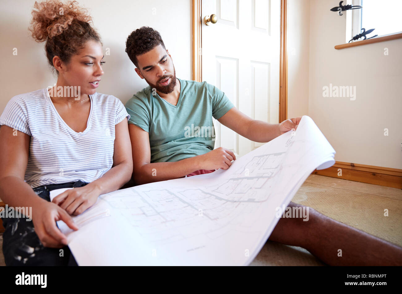 Couple Sitting On Floor Looking At Plans In Empty Room Of New Home