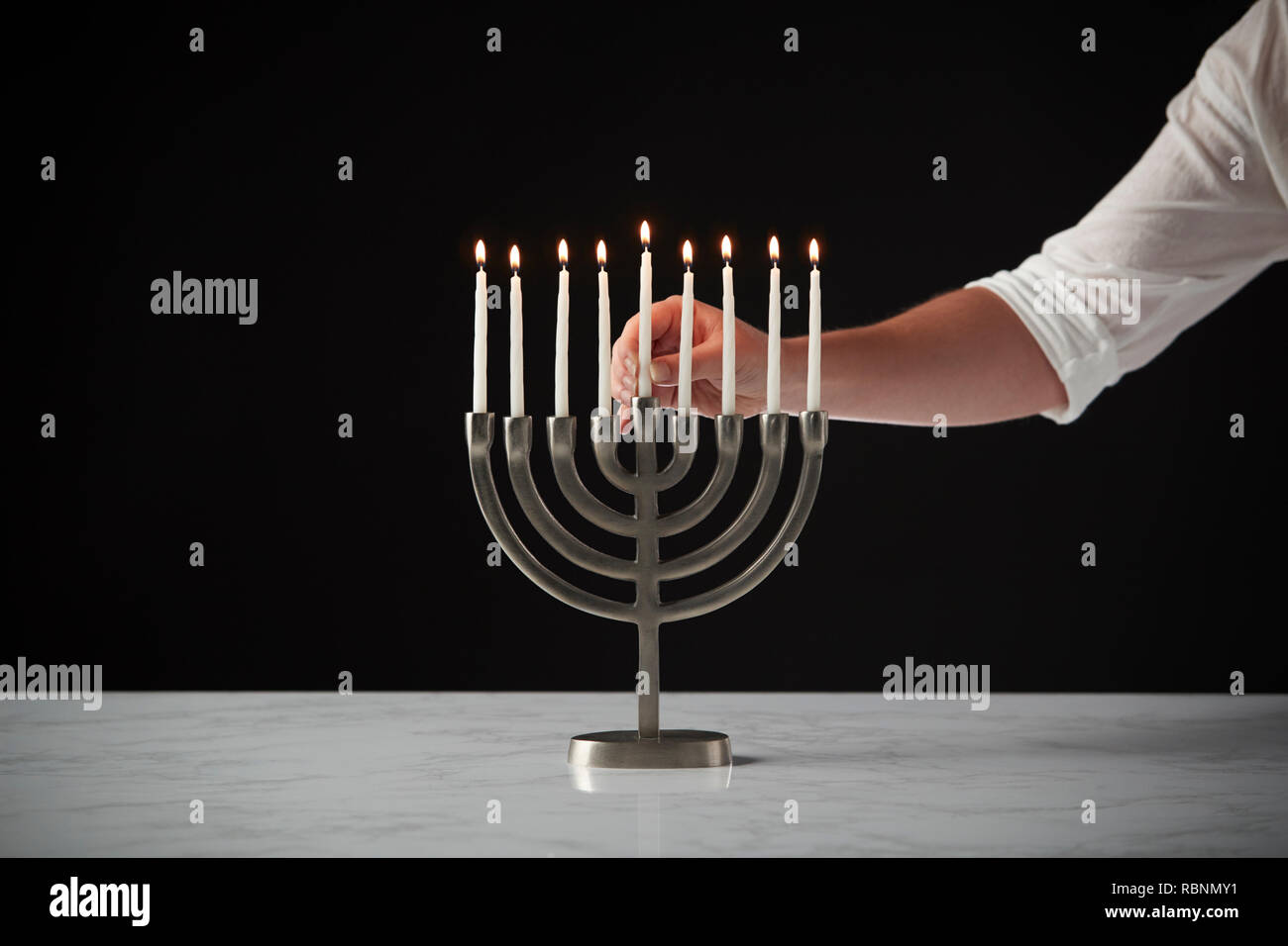 Hand Placing Lit Candle On Metal Hanukkah Menorah On Marble Surface Against Black Studio Background Stock Photo