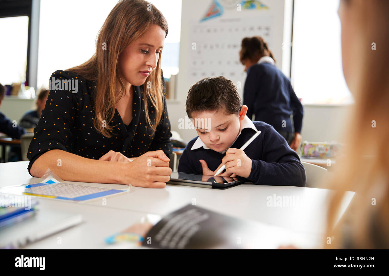 Young female teacher working with a Down syndrome schoolboy sitting at desk using a tablet computer and stylus in a primary school classroom, selective focus Stock Photo