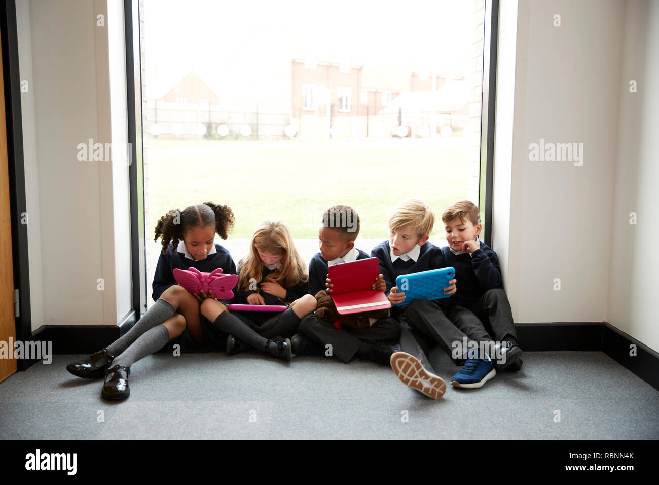 Five primary school kids sitting in a row on the floor in front of a window in a school corridor looking at tablet computers, front view, close up Stock Photo