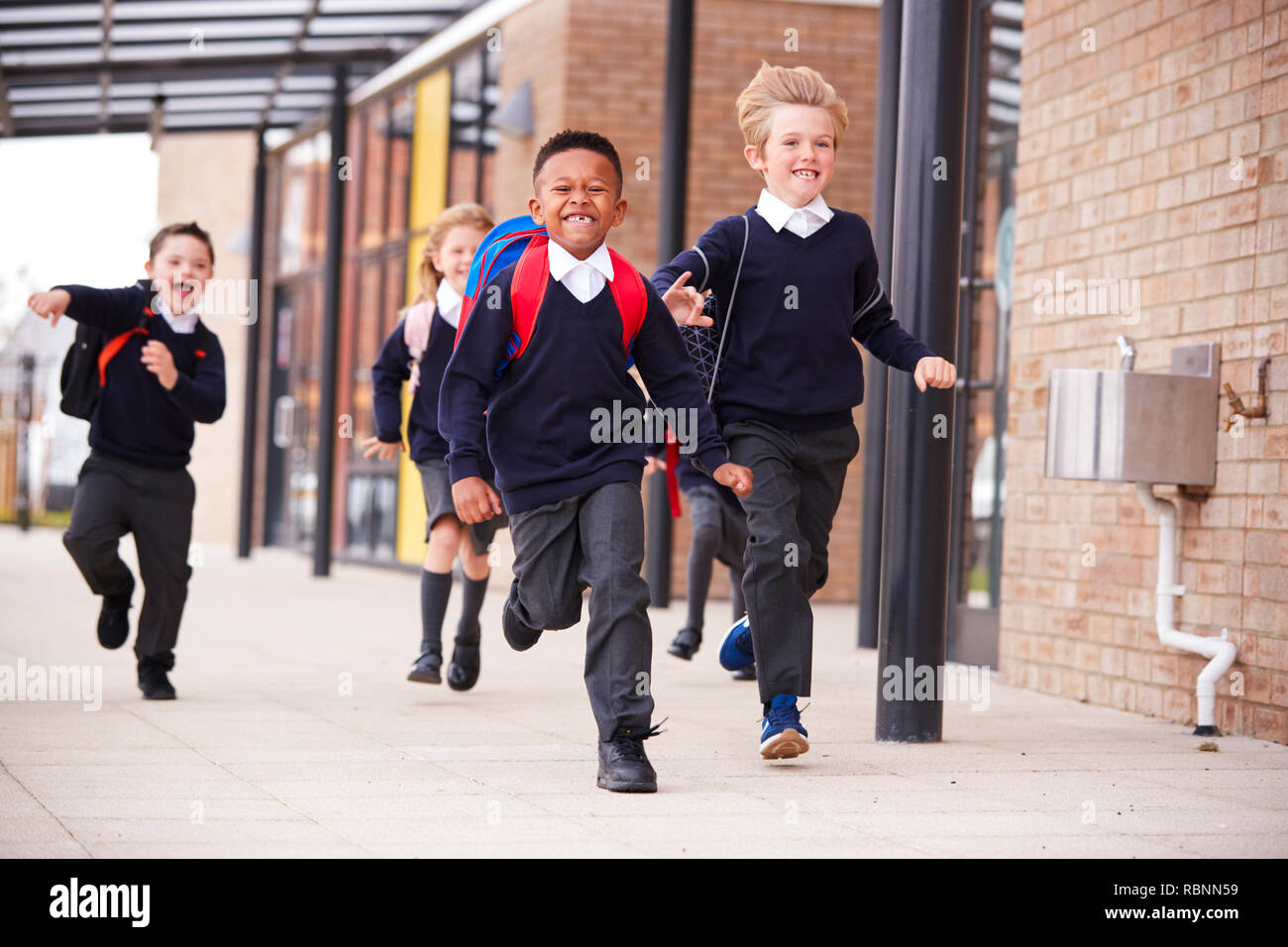 Happy primary school kids, wearing school uniforms and backpacks, running on a walkway outside their school building, front view, close up Stock Photo