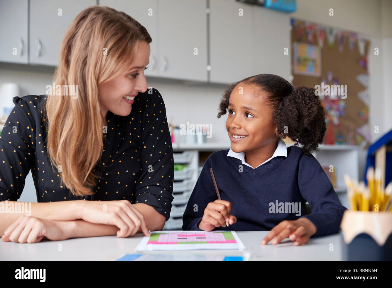 Young female primary school teacher working one on one with a schoolgirl at a table in a classroom, both looking at each other smiling, close up Stock Photo