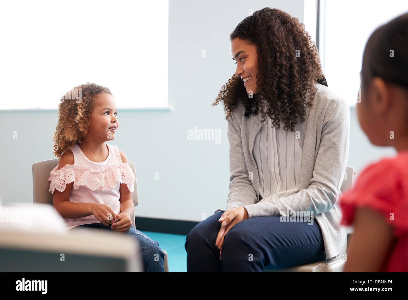 Infant school girl and her female teacher sitting on chairs in a classroom smiling to each other, close up Stock Photo