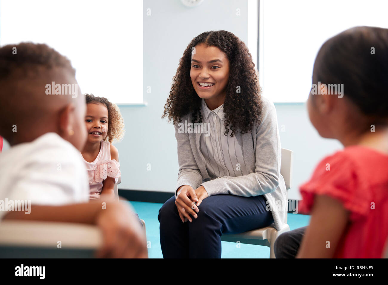 Infant school children sitting on chairs in a circle in a classroom talking to their smiling female teacher, close up, selective focus Stock Photo