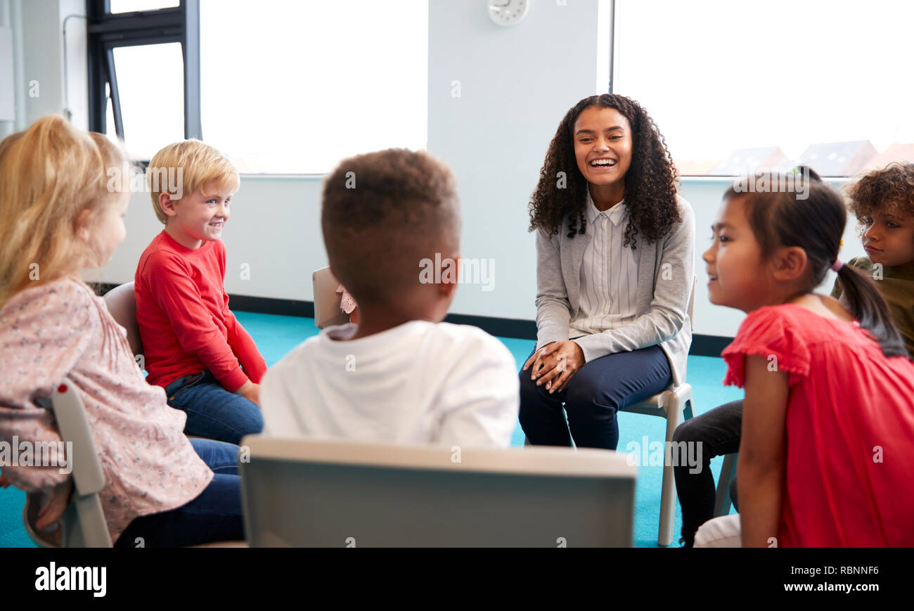 A class of infant school children sitting on chairs in a circle in the classroom talking to their female teacher Stock Photo
