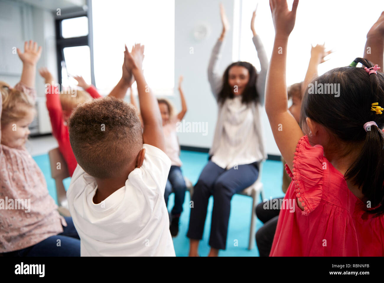 A class of infant school children sitting on chairs in a circle in the classroom, raising hands with their female teacher, close up Stock Photo