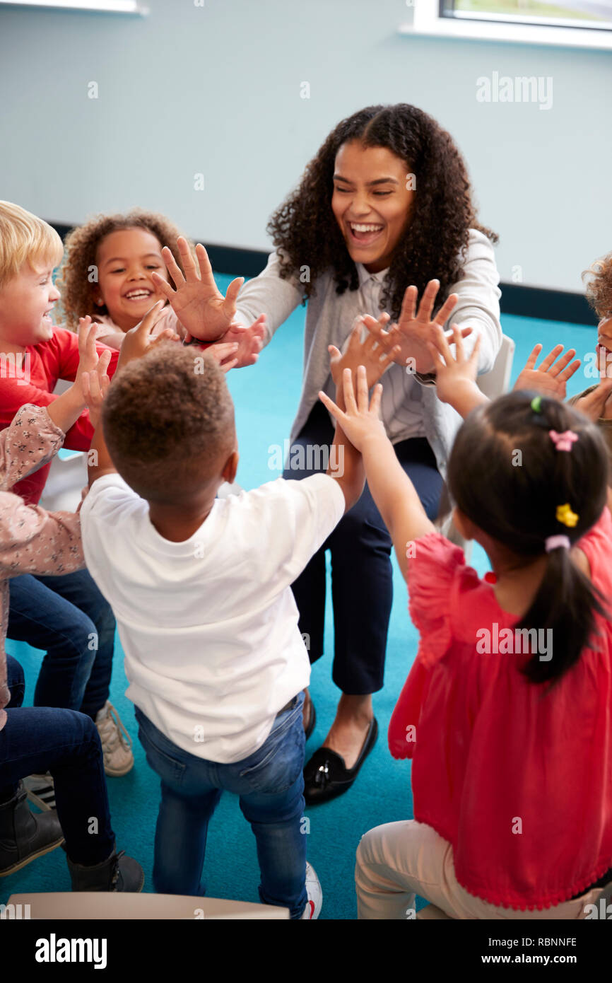 Elevated view of infant school children in a circle in the classroom giving high fives to their smiling female teacher, vertical, close up Stock Photo