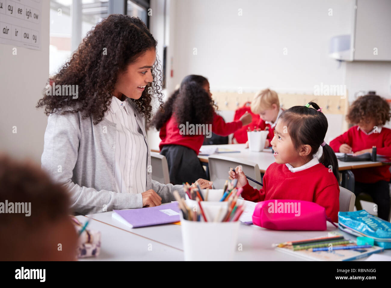 Female kindergarten teacher sitting at table in a classroom talking to a young Chinese schoolgirl, close up Stock Photo