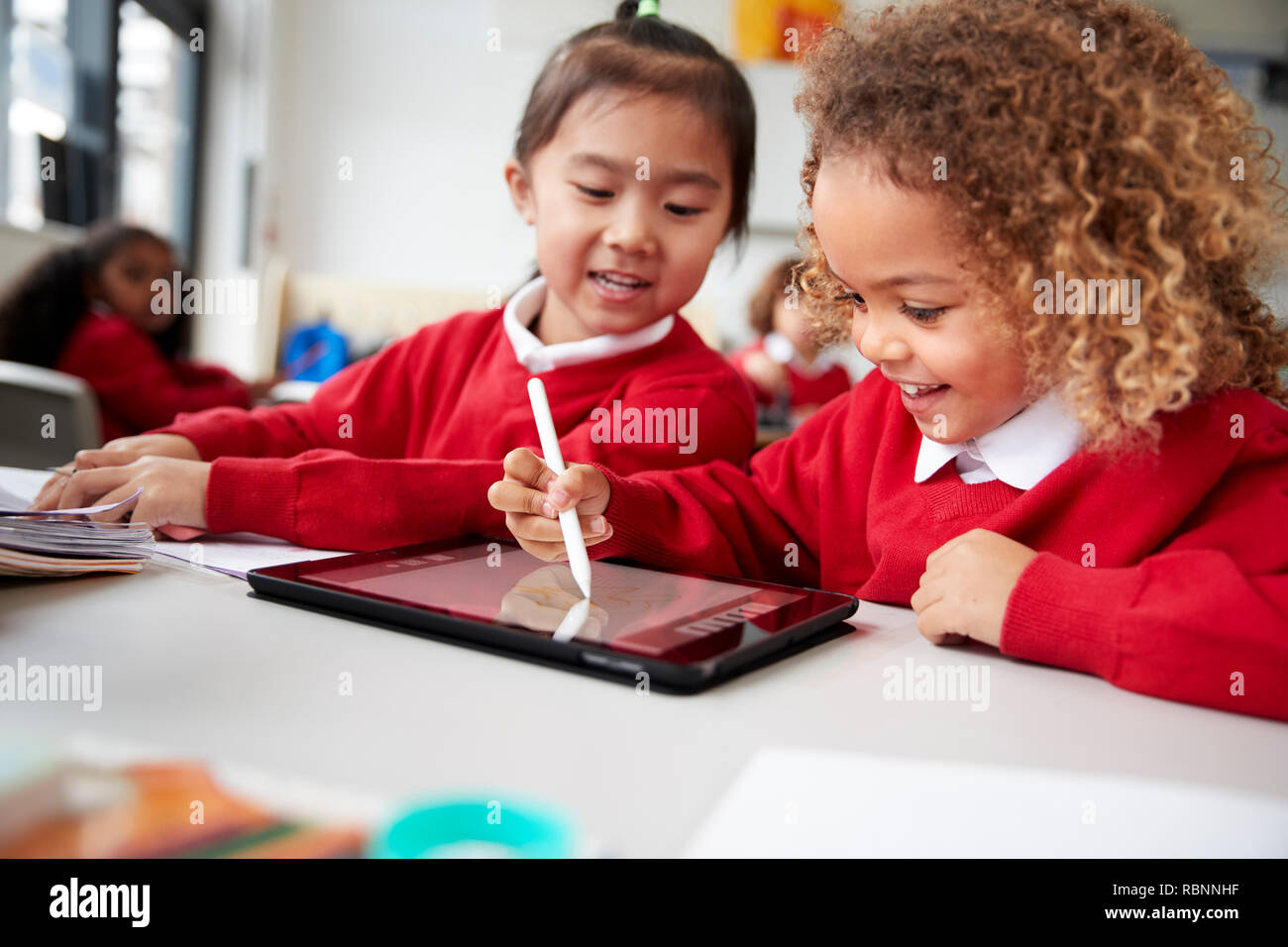 Close up of two kindergarten schoolgirls wearing school uniforms, sitting at a desk in a classroom using a tablet computer and stylus, looking at screen and smiling Stock Photo