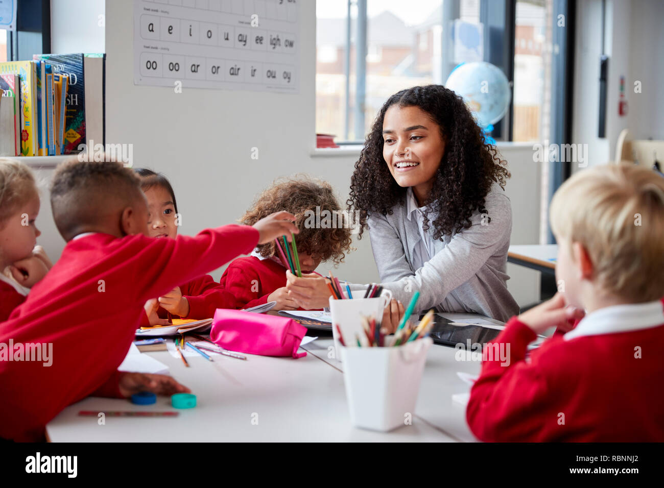 Front view of smiling female infant school teacher sitting at a table in the classroom with a group of schoolchildren Stock Photo