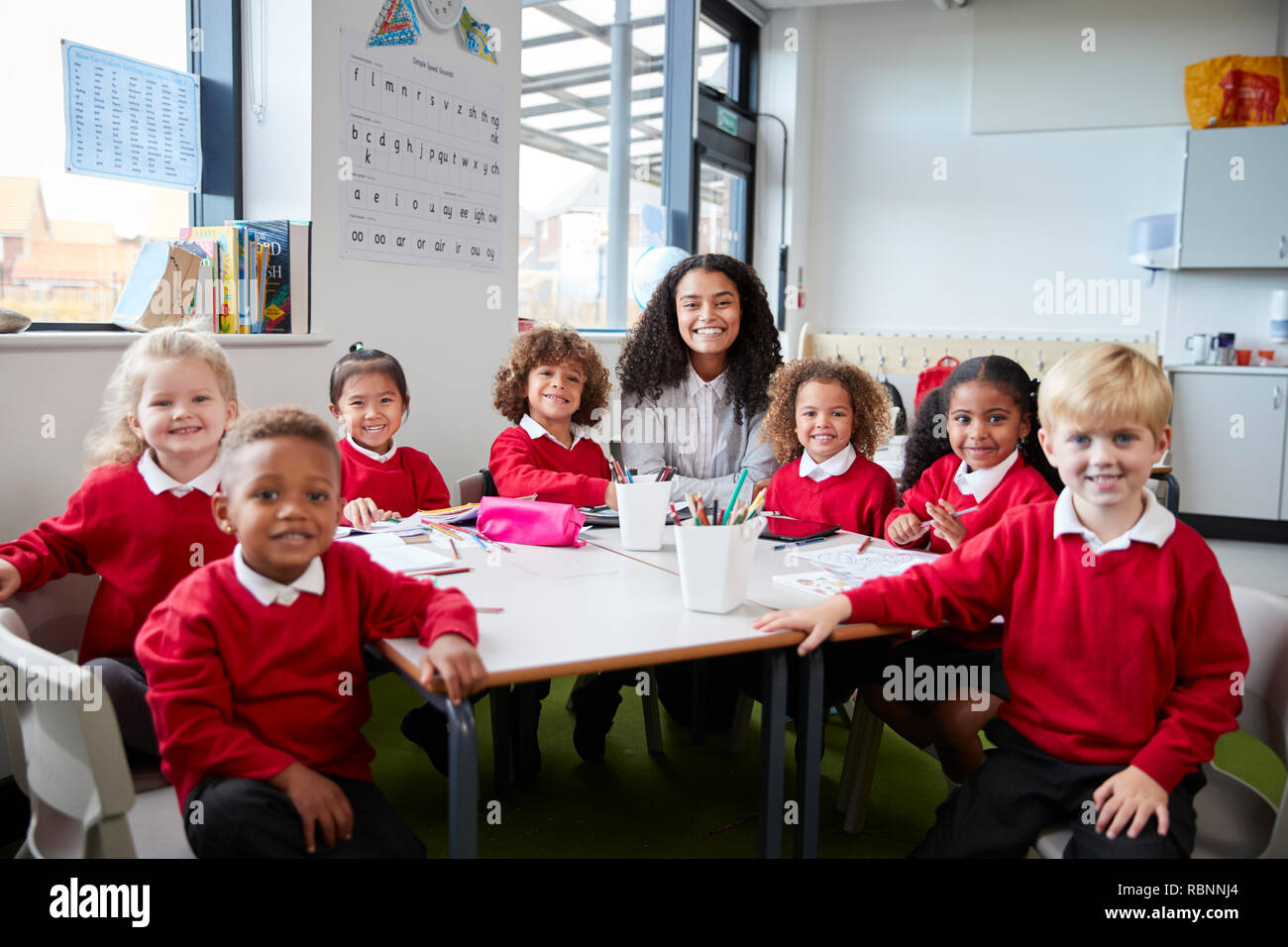 Group portrait of infant school teacher and kids sitting at table in a classroom looking to camera smiling, front view Stock Photo