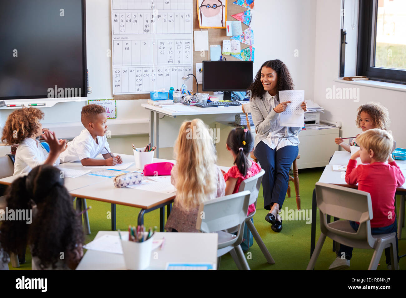 Smiling female infant school teacher sitting on a chair facing school kids in a classroom holding up and explaining a worksheet to them Stock Photo