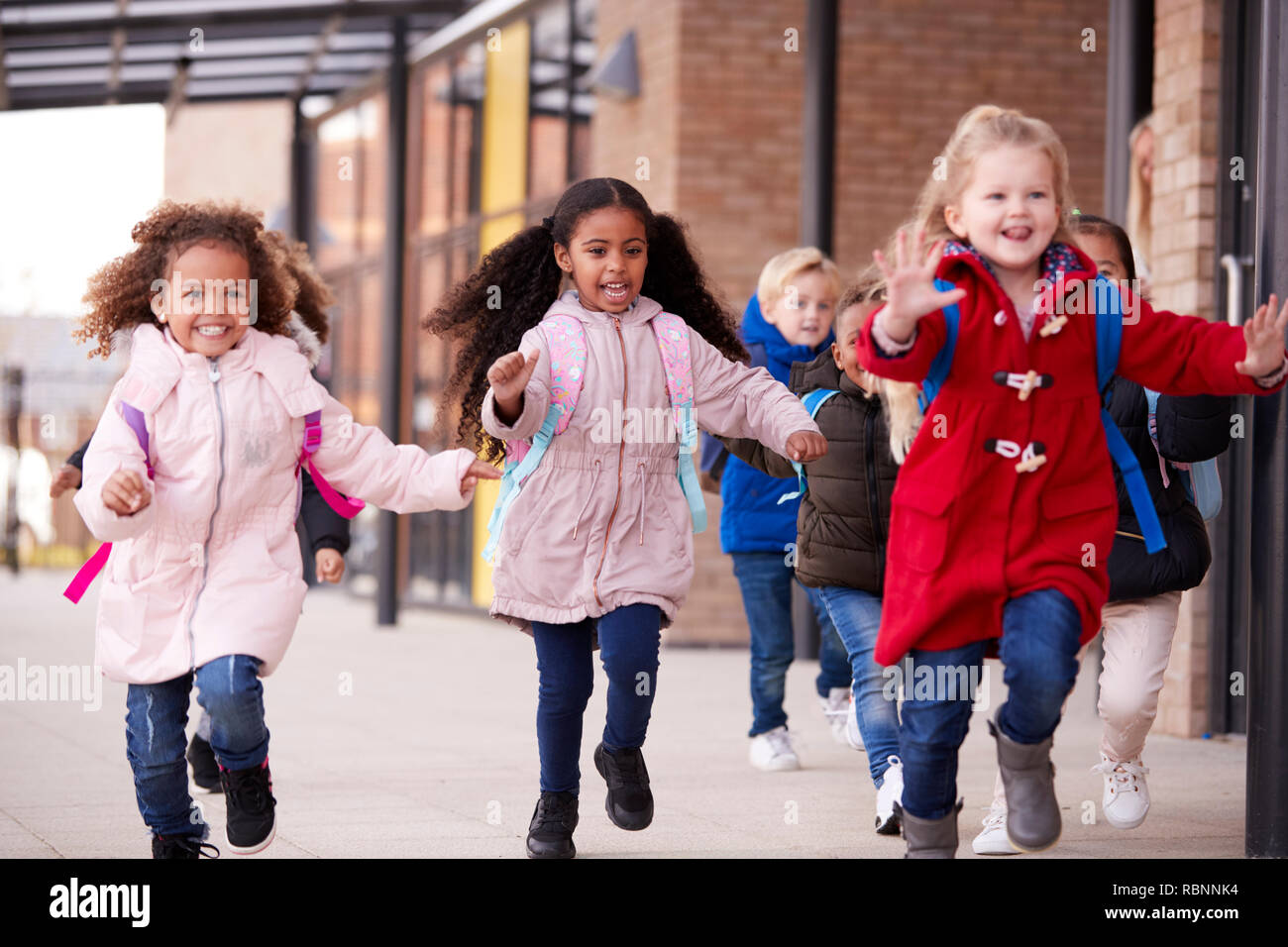 Three happy young school girls wearing coats and carrying schoolbags running in a walkway with their classmates outside their infant school building, close up Stock Photo