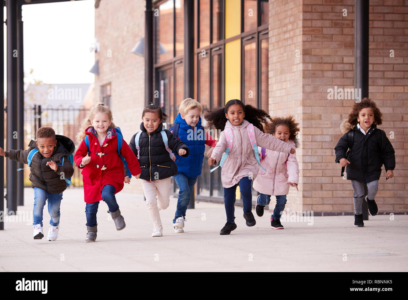 A happy multi-ethnic group of young school kids wearing coats and carrying schoolbags running in a walkway with their classmates outside their infant school building Stock Photo