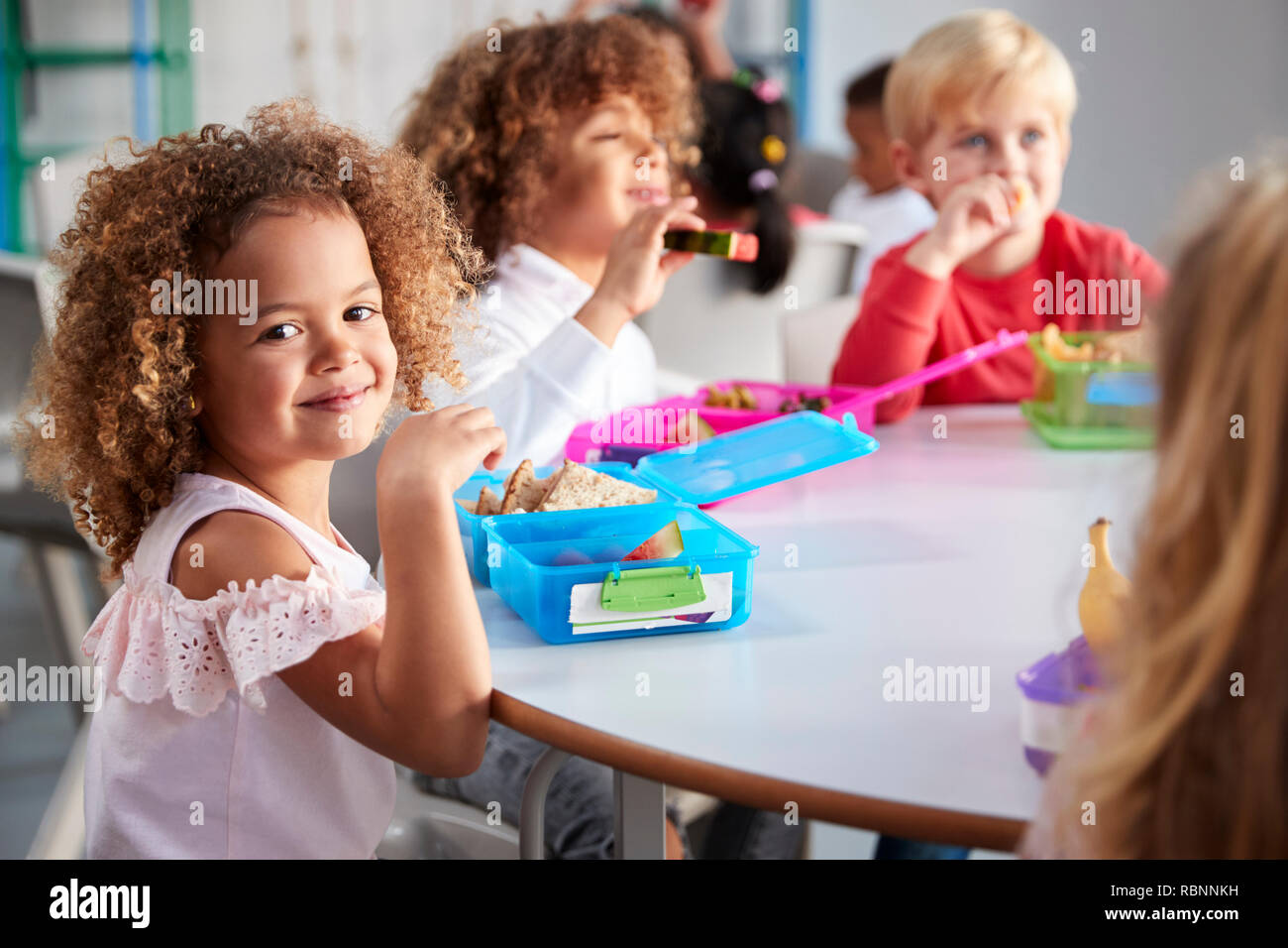 Close up of smiling young children sitting at a table eating their packed lunches together at infant school, girl smiling to camera, selective focus Stock Photo