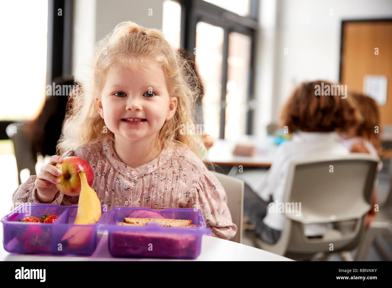 Young white schoolgirl sitting at a table smiling and holding an apple in a kindergarten classroom during her lunch break, close up Stock Photo
