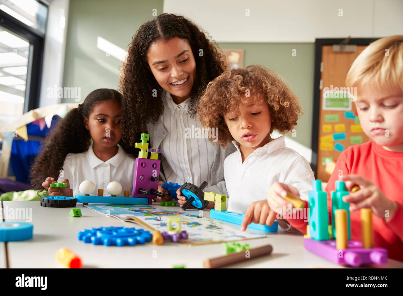 Female teacher sitting at table in play room with three kindergartne children constructing, selective focus Stock Photo