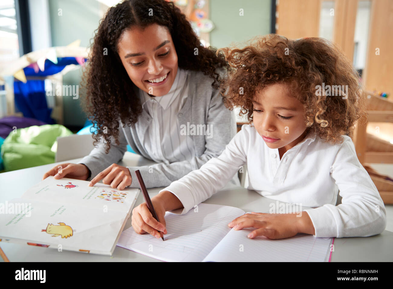 Female infant school teacher working one on one with a young schoolgirl sitting at a table writing in a classroom, front view, close up Stock Photo