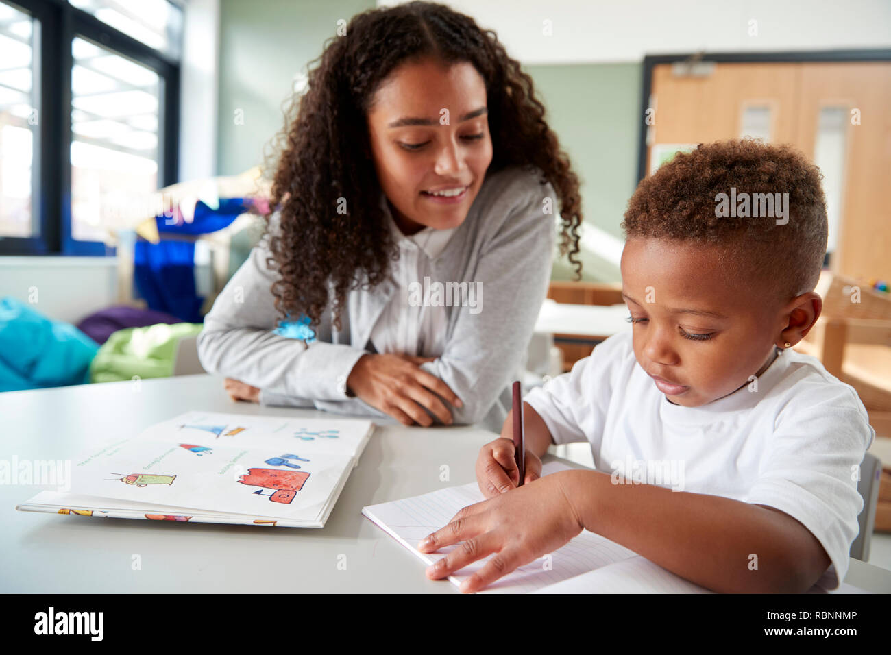 Female infant school teacher working one on one with a young schoolboy, sitting at a table writing in a classroom, front view, close up Stock Photo