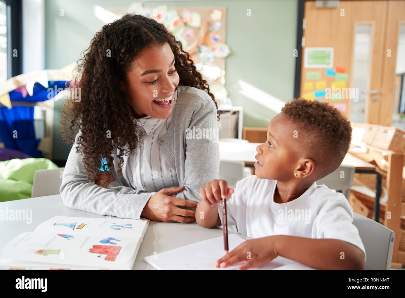 Female infant school teacher working one on one with a young schoolboy, sitting at a table smiling at each other, close up Stock Photo