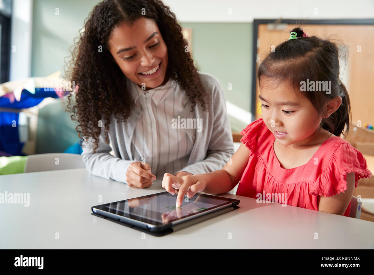 Female infant school teacher working one on one with a Chinese schoolgirl, sitting at a table in a classroom using a tablet computer, close up Stock Photo