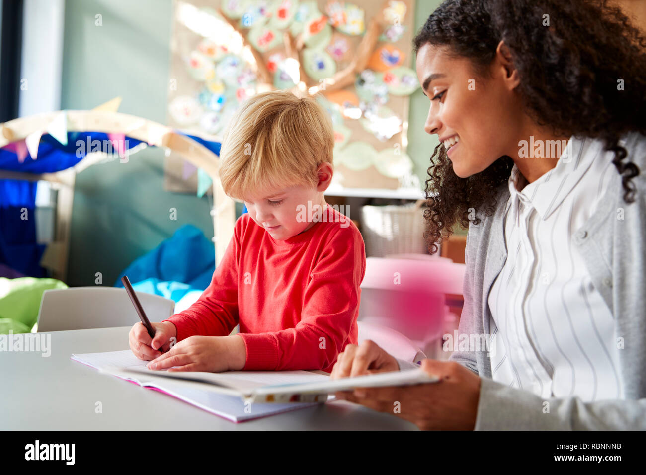 Female infant school teacher working one on one with a young white schoolboy, sitting at a table in a classroom writing, close up Stock Photo